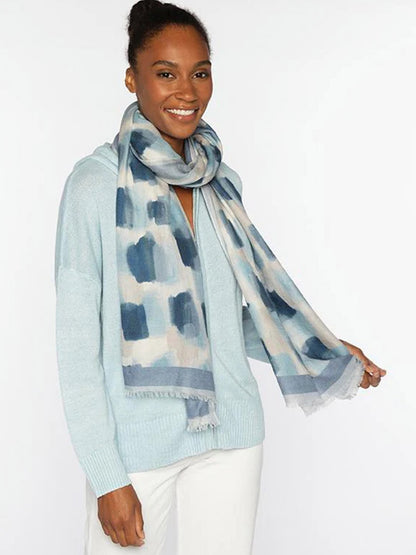 A woman smiles against a plain white background, wearing a light blue sweater, white pants, and the Kinross Brushstroke Print Scarf in Ink Multi.