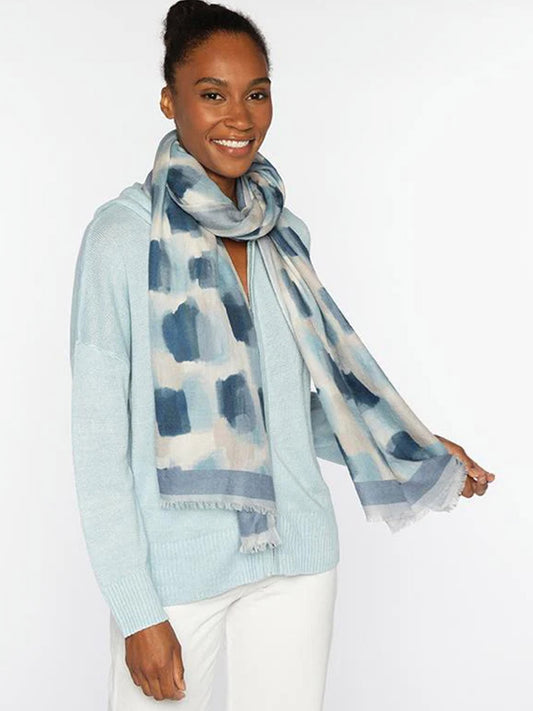 A woman smiles against a plain white background, wearing a light blue sweater, white pants, and the Kinross Brushstroke Print Scarf in Ink Multi.