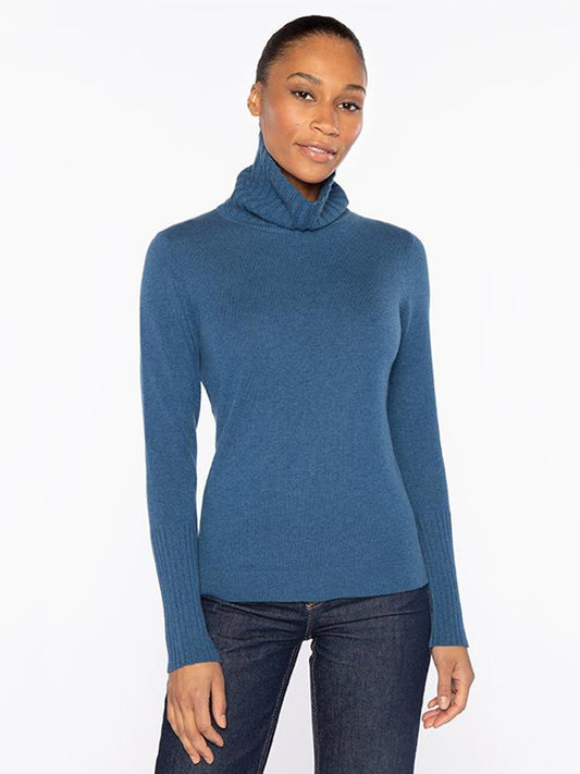 A woman stands against a plain background wearing the Kinross Chunky Trim T-Neck in Sapphire with dark jeans, her hair pulled back and arms relaxed at her sides.