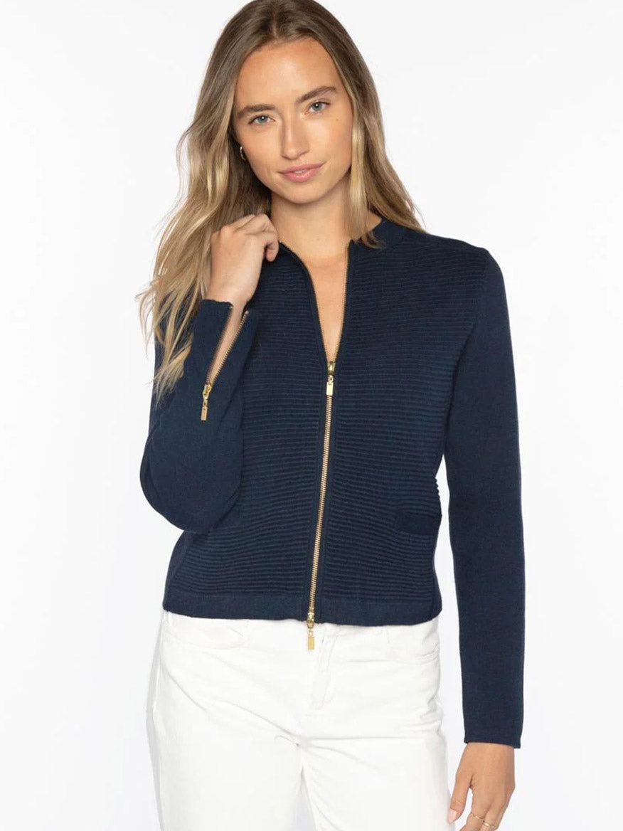 A woman stands against a plain background in the Kinross Fitted Zip Cardigan in Navy, made from SuperSoft Cotton, paired with white pants. Her left hand touches her hair, showcasing a modern approach to women's knitwear.