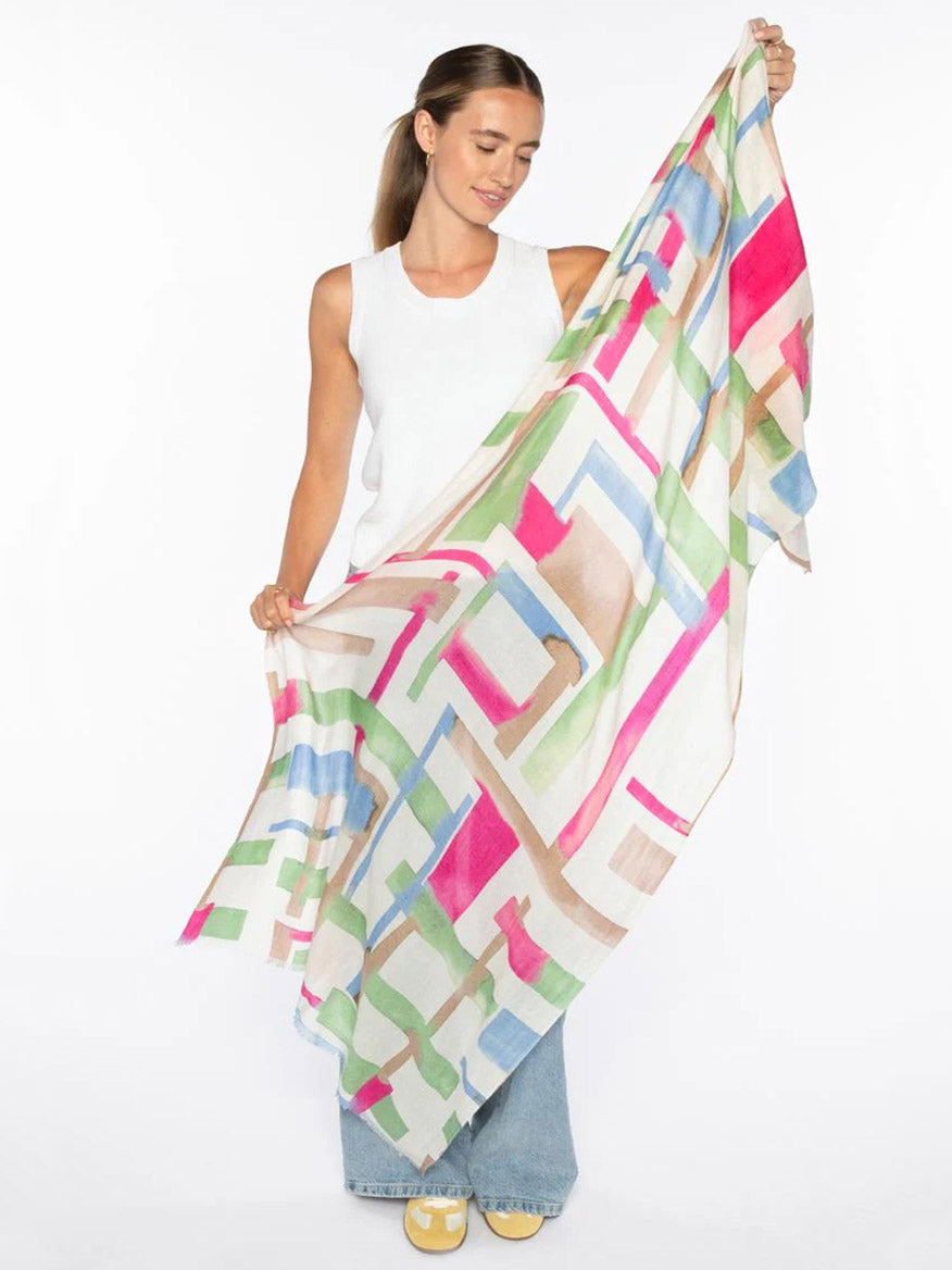 A woman in a white sleeveless top and blue jeans holds the Kinross Geo Print Scarf in Dahlia Multi, displayed against a white background.