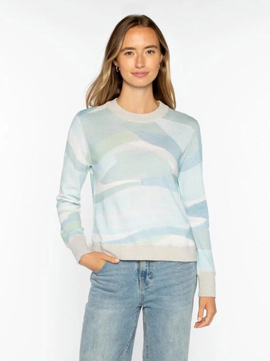 A woman wears the Kinross Landscape Print Pullover in Multi, a pastel cotton-cashmere blend sweater with abstract patterns, paired with light blue jeans, standing against a plain white background.