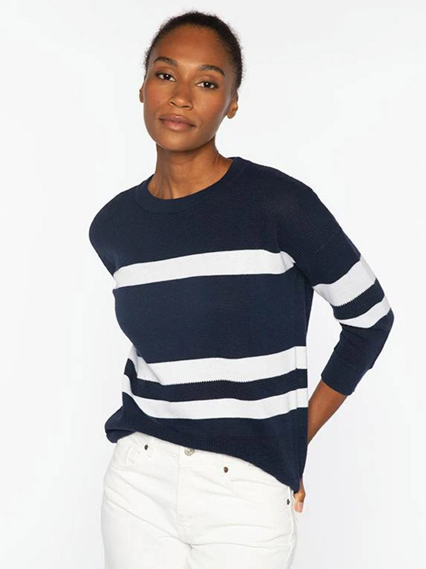 A woman wears the Kinross Mesh Stripe Crew in Denim/Greystone— a navy blue pullover with white stripes—paired with white pants, standing against a plain white background.