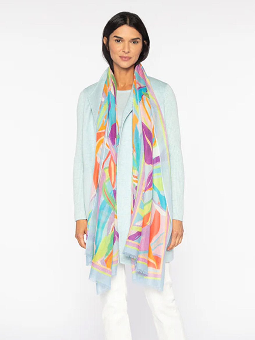 A person stands against a plain background wearing the Kinross Palm Print Scarf in Freesia over a light blue sweater and white pants.