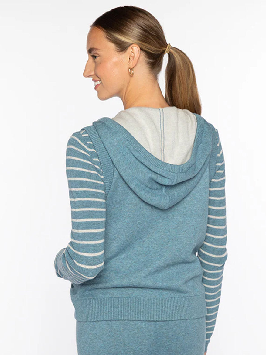 A woman with a ponytail stands facing away against a white background, wearing the Kinross Sleeveless Zip Hoodie in Denim with striped sleeves.
