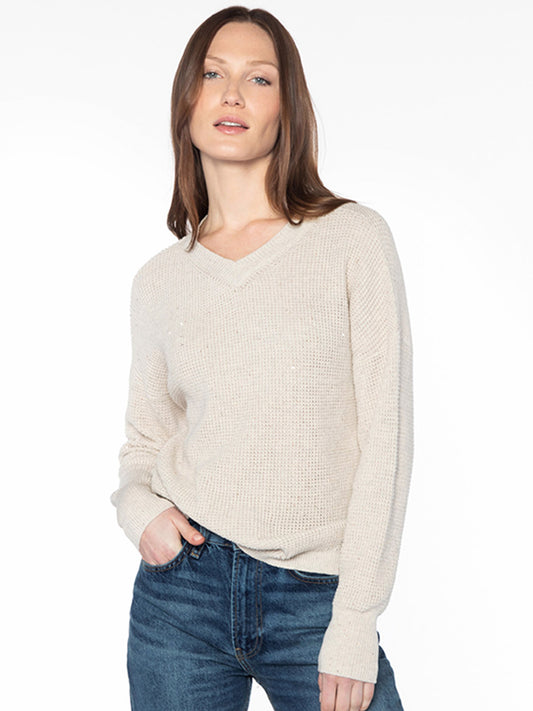 A person with long hair wears the Kinross Thermal Sequin Vee in Champagne along with blue jeans, standing against a plain white background.