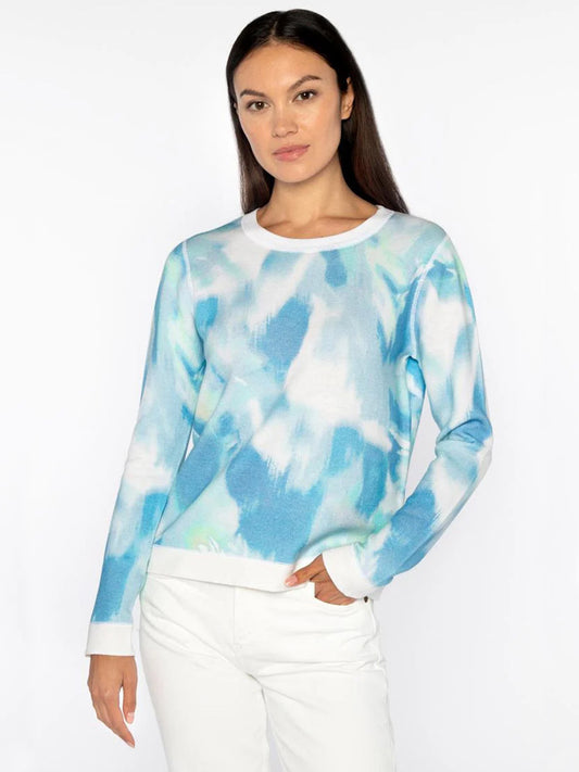 A person stands against a plain background, wearing the Kinross Waterway Print Crew in Multi, made from a blue and white tie-dye cotton cashmere blend sweater, paired with crisp white pants.