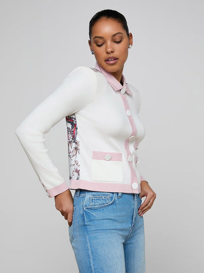 A woman stands against a gray background wearing the L'Agence Nomi Satin Back Cardigan in Ivory/Pink/Multi Butterfly Jungle, featuring contrast trim and statement buttons, paired with light blue jeans.