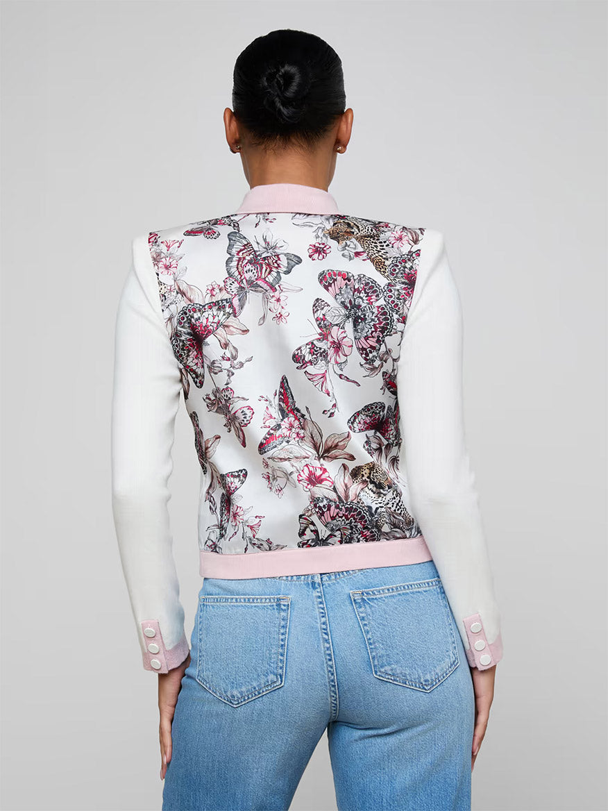 A person faces away, wearing light blue jeans and the L'Agence Nomi Satin Back Cardigan in Ivory/Pink/Multi Butterfly Jungle, featuring a butterfly print and statement buttons on the back.
