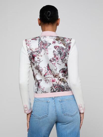 A person faces away, wearing light blue jeans and the L'Agence Nomi Satin Back Cardigan in Ivory/Pink/Multi Butterfly Jungle, featuring a butterfly print and statement buttons on the back.