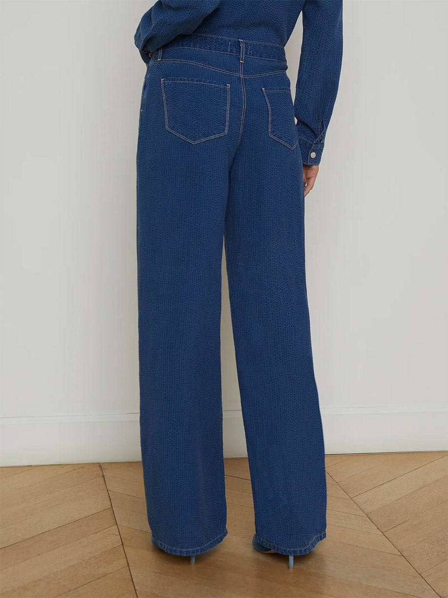 A person stands with their back to the camera on a wood floor, wearing L'Agence Alicent Wide-Leg Jean in Heron and a matching long-sleeve shirt against a plain wall.