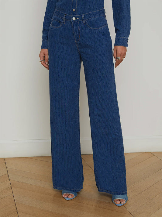 A person stands on a wooden floor wearing the L'Agence Alicent Wide-Leg Jean in Heron, a long-sleeve blue shirt, and blue open-toe heels. The jeans are high-rise and sneaker length. The person's head is not visible in the image.