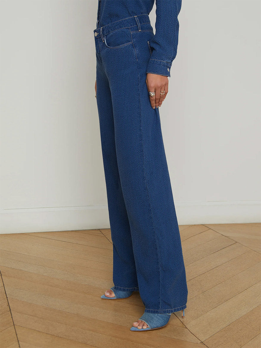 Indoors on a wooden floor, someone wears the L'Agence Alicent Wide-Leg Jean in Heron, a matching seersucker chambray shirt, and open-toed heeled shoes.