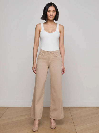 Woman standing indoors against a plain wall, wearing a white sleeveless top, the L'Agence Alicent Wide-Leg Jean in Pale Khaki, and beige heels.