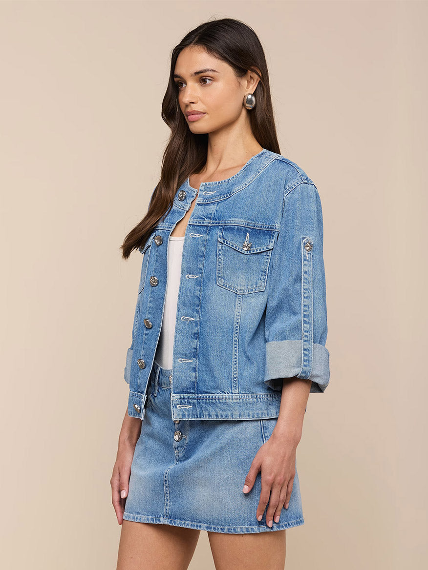 A woman wears the L'Agence Anouk Jean Jacket in Grant, a light blue all-cotton denim jacket with silver buttons, paired with a matching skirt, standing against a plain beige background.