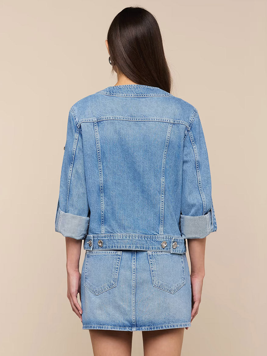 A woman with long brown hair models the L'Agence Anouk Jean Jacket in Grant, an indigo cotton denim piece with rolled sleeves, paired with a matching mini skirt, shown from the back against a beige background.