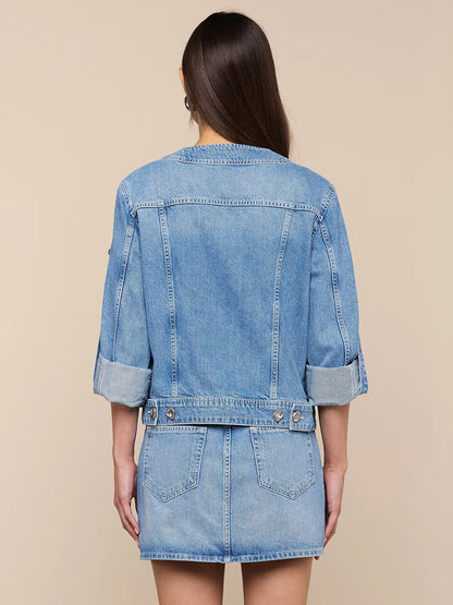 A woman with long brown hair models the L'Agence Anouk Jean Jacket in Grant, an indigo cotton denim piece with rolled sleeves, paired with a matching mini skirt, shown from the back against a beige background.