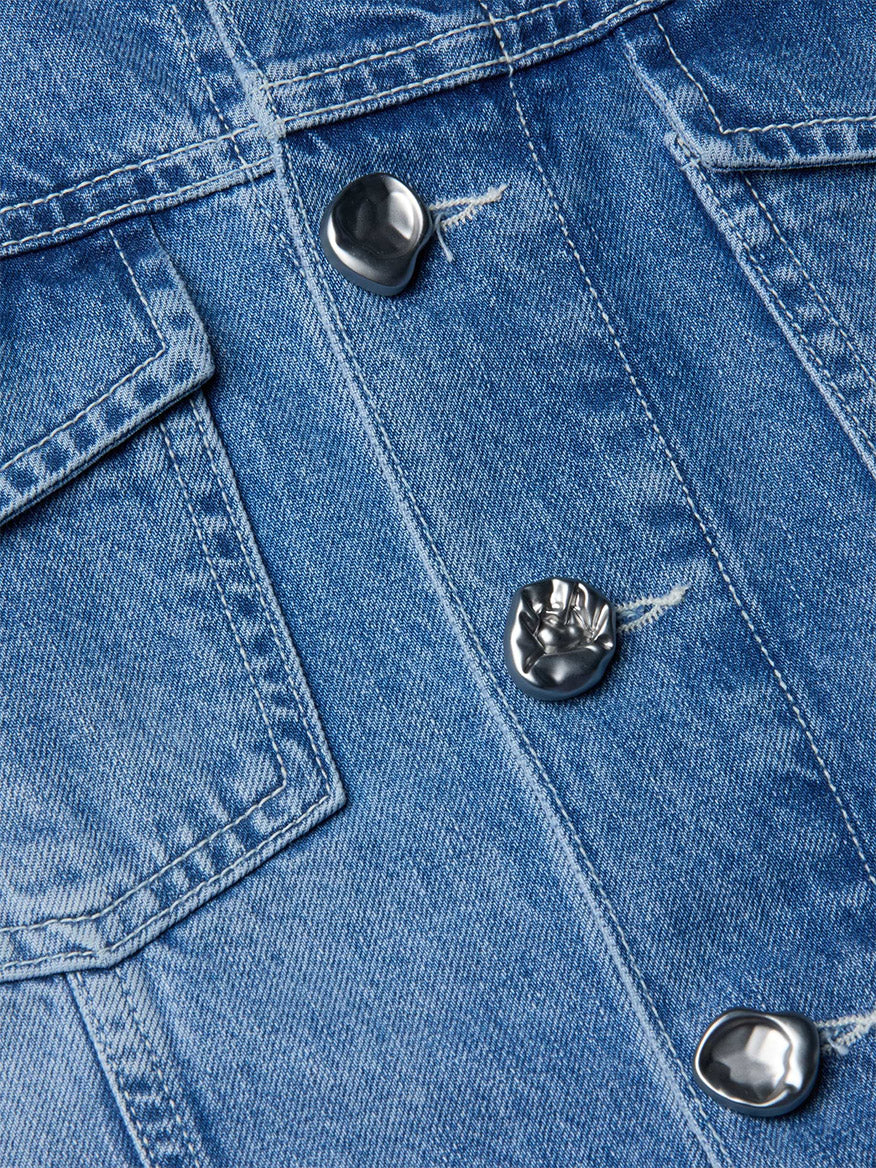 Close-up of the L'Agence Anouk Jean Jacket in Grant, a collarless blue indigo denim piece with silver irregular metal buttons and visible stitching by the chest pocket.