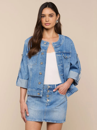 A woman wears the L'Agence Anouk Jean Jacket in Grant with a matching all-cotton denim skirt and a white top, standing against a plain beige background.