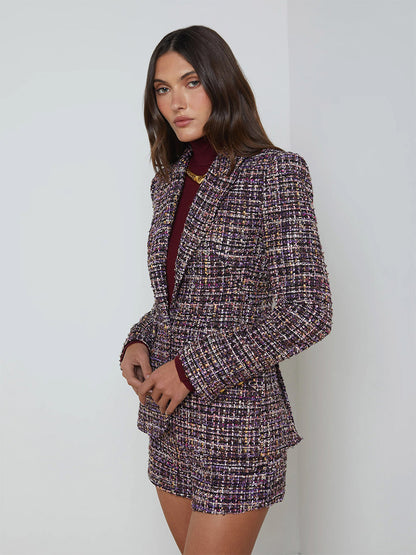 A woman wearing the L'Agence Chamberlain Tweed Blazer in Purple Multi and shorts stands against a white wall, looking at the camera.