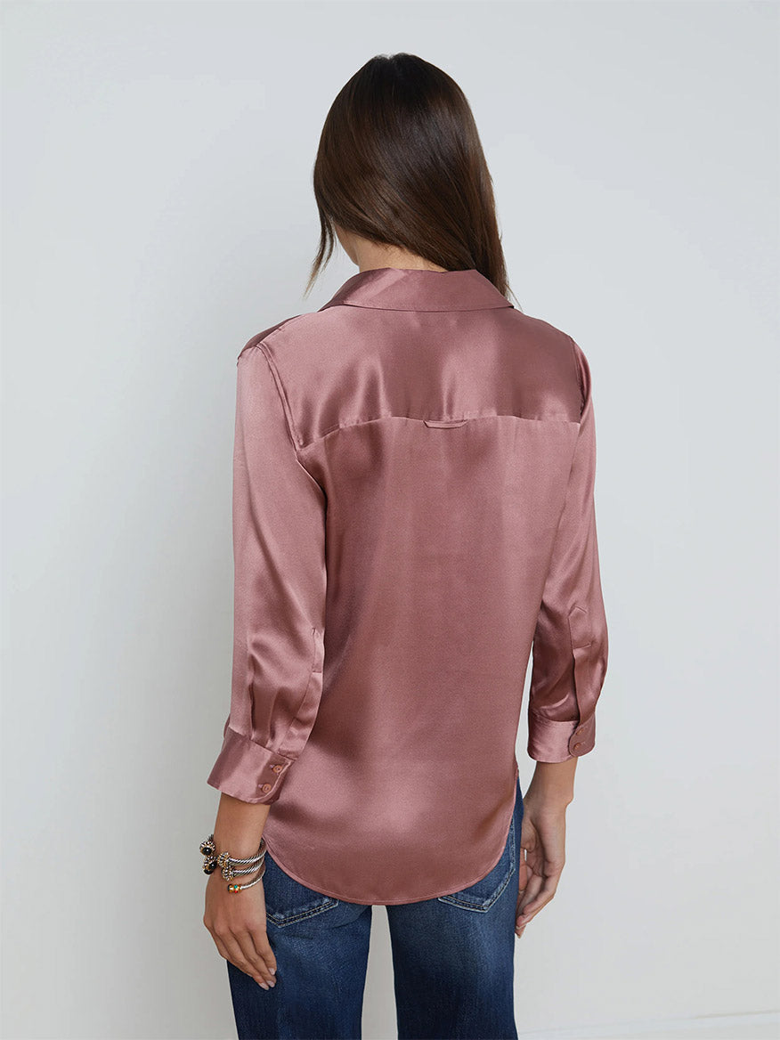 A person with long hair, seen from behind, wearing a L'Agence Dani Blouse in Mauve Rose and blue jeans stands against a plain white background, complemented by a stylish single-breasted blazer with embossed dome buttons.