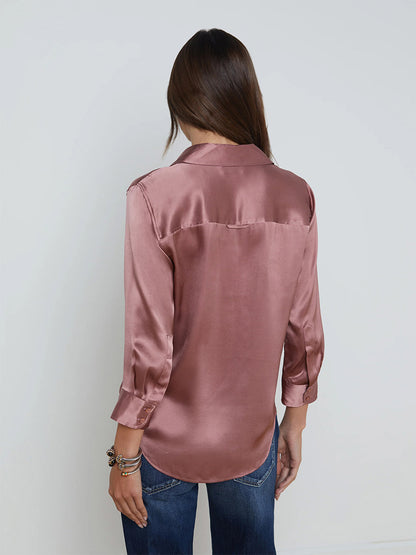 A person with long hair, seen from behind, wearing a L'Agence Dani Blouse in Mauve Rose and blue jeans stands against a plain white background, complemented by a stylish single-breasted blazer with embossed dome buttons.