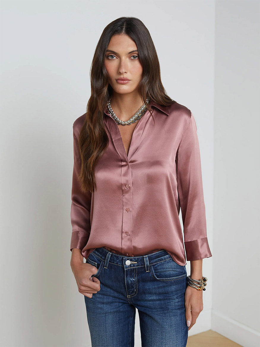 A woman with long brown hair, wearing the L'Agence Dani Blouse in Mauve Rose and dark blue jeans, stands in a room with a white wall and wooden floor. Over her blouse, she dons a chic single-breasted blazer featuring embossed dome buttons, adding an elegant touch to her casual outfit.