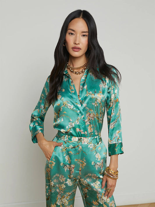 A person stands wearing a L'Agence Dani Blouse in Teal Multi Watercolor Cherry Blossom with matching pants, accessorized with layered gold necklaces and bracelets, against a plain background reminiscent of a NYFW presentation.