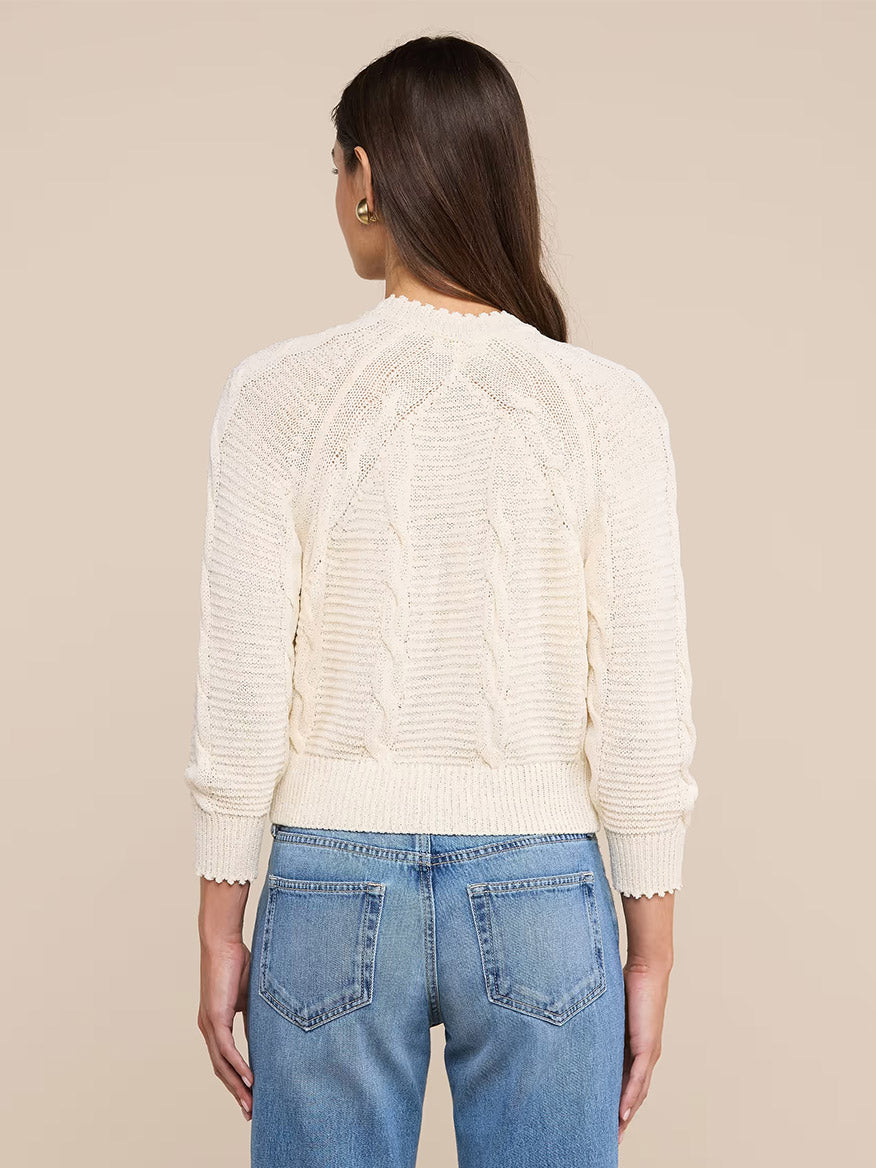 A woman with long brown hair faces away, wearing the L'Agence Darcy Cable Knit Cardigan in Ivory, featuring button pockets, paired with light blue jeans against a neutral background.