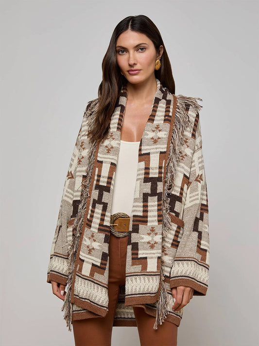 A woman faces forward against a plain background, wearing the L'Agence Jair Cardigan in Natural Multi with fringe trim, a light top, brown pants, and gold earrings.
