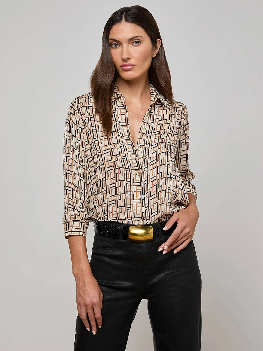 A woman with straight brown hair wears the L'Agence Nina Silk Blouse in Ecru Multi Geo Print, tucked into black pants with a wide belt and large gold buckle. She stands against a plain light background.
