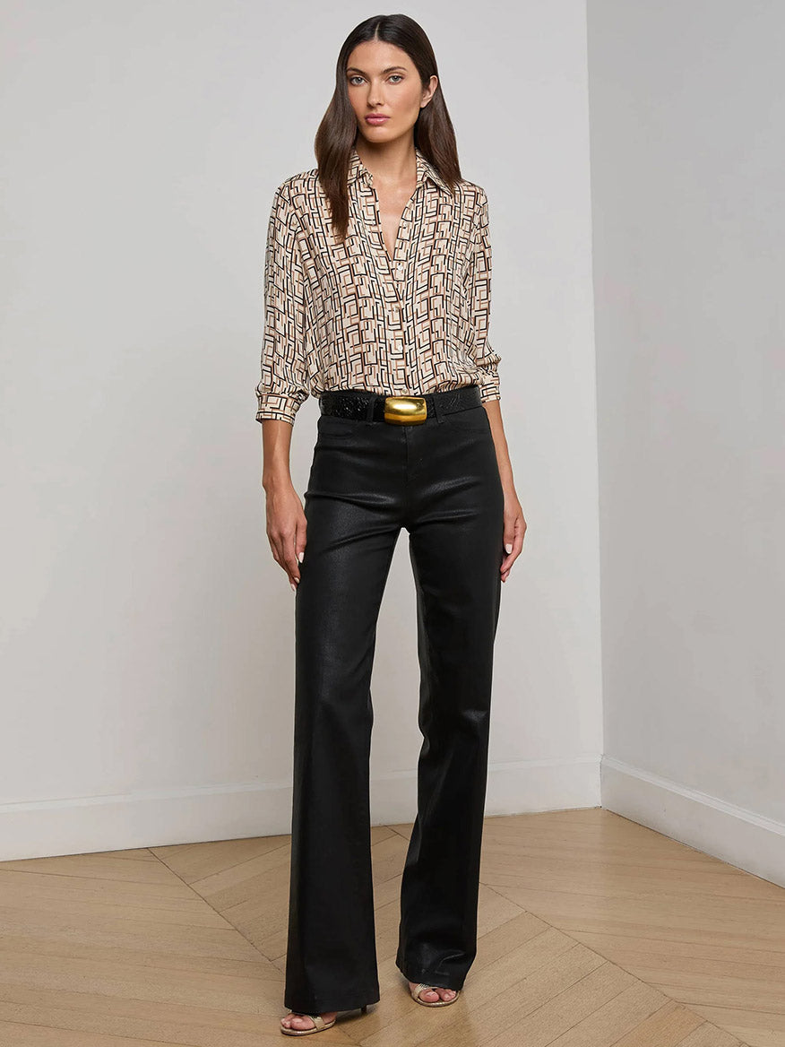 A woman stands indoors on a wooden floor wearing the L'Agence Nina Silk Blouse in Ecru Multi Geo Print, tucked into high-waisted black pants with a gold belt buckle and open-toed heels.