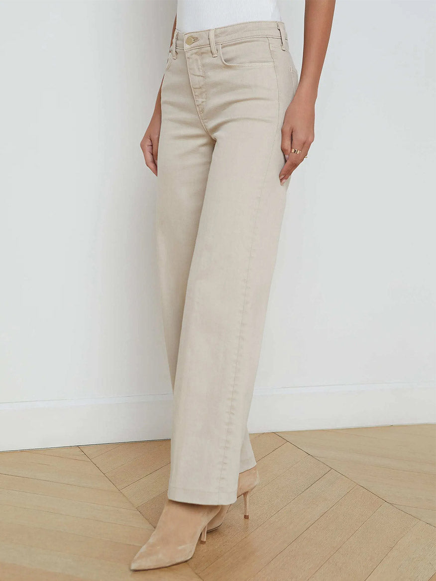 A person wearing L'Agence Scottie Wide-Leg Jean in Biscuit with nude heels stands on a wooden floor against a plain white wall.
