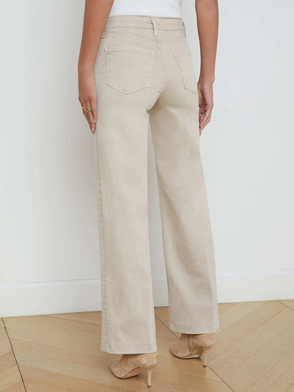 A person stands with their back to the camera against a white wall, wearing L'Agence Scottie Wide-Leg Jean in Biscuit—high-rise, wide-leg beige jeans made from premium stretch denim—and nude heels.
