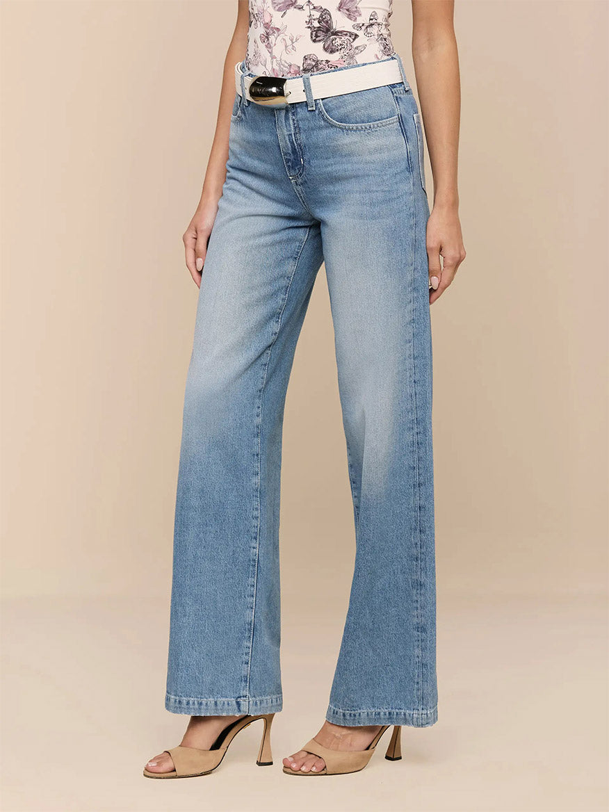 Wearing L'Agence Scottie Wide-Leg Jean in Grant, a white belt, a butterfly-print top, and beige heeled sandals, the person stands against a plain beige background.