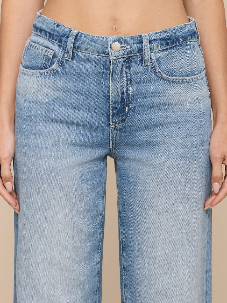 Close-up of a person wearing the L'Agence Scottie Wide-Leg Jean in Grant, a high-rise, light blue all-cotton denim style with button and zipper front, standing against a neutral background.