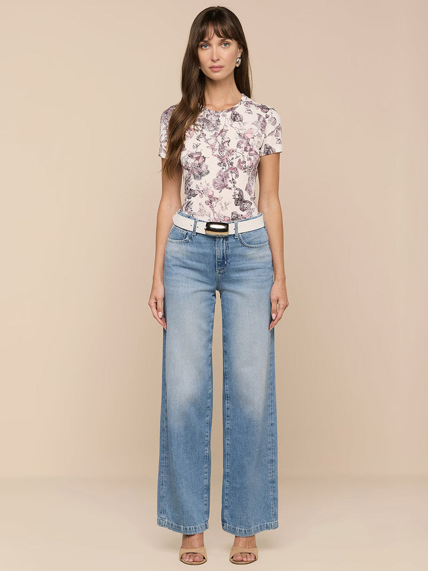 A woman stands against a plain background wearing a floral print t-shirt, the L'Agence Scottie Wide-Leg Jean in Grant, a white belt, and heeled sandals.
