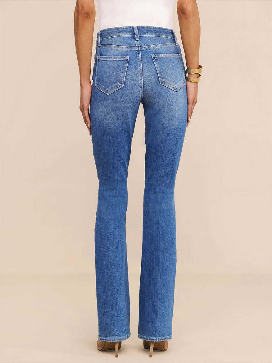 A person is shown from the back against a plain light background, wearing the L'Agence Selma Bootcut Jean in Sullivan—a high-waisted, medium wash bootcut style—paired with heeled shoes.