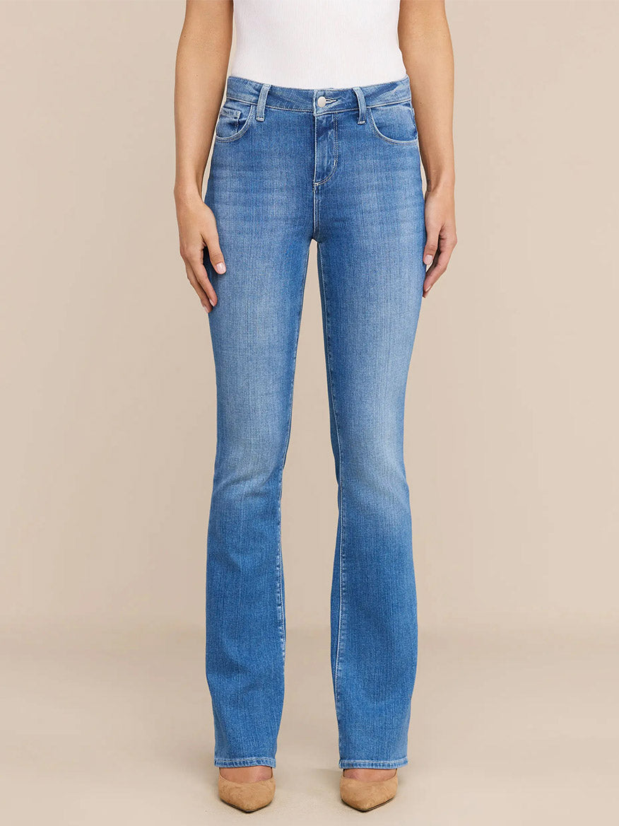 Wearing the L'Agence Selma Bootcut Jean in Sullivan, a white sleeveless top, and tan shoes, the person stands against a plain beige background.