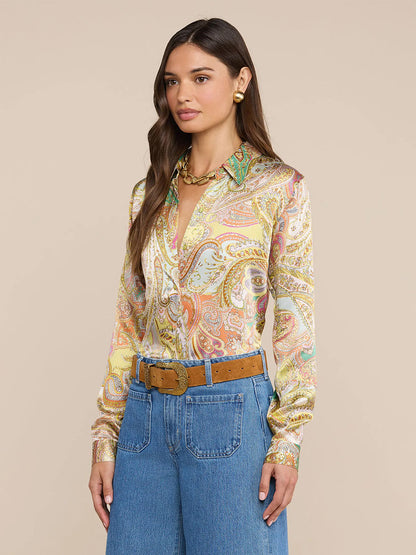Woman wearing the L'Agence Tyler Blouse in Green Multi Cluster Paisley, paired with blue high-waisted jeans, a brown belt, gold earrings, and a gold necklace, standing against a plain beige background.