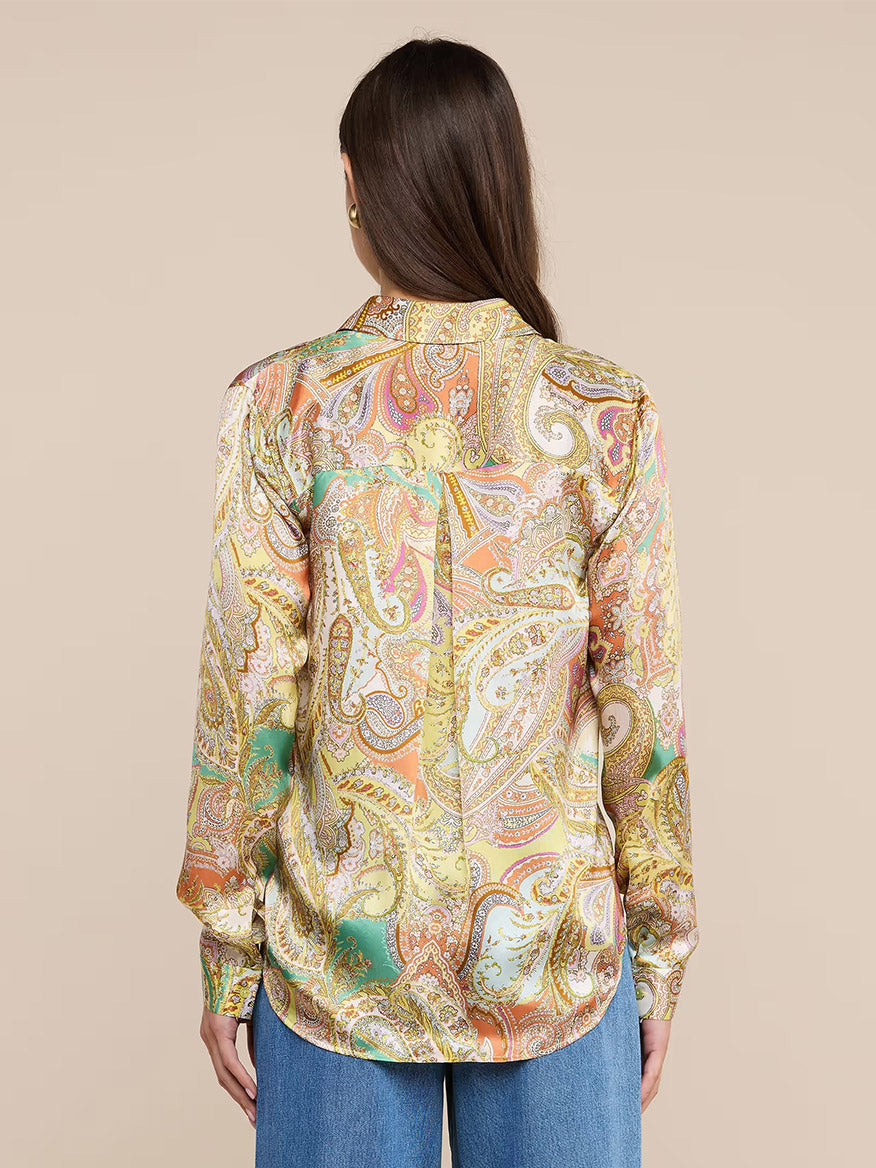 A woman with long brown hair is seen from the back against a plain beige background, wearing blue jeans and the L'Agence Tyler Blouse in Green Multi Cluster Paisley, a colorful silk button-down shirt.