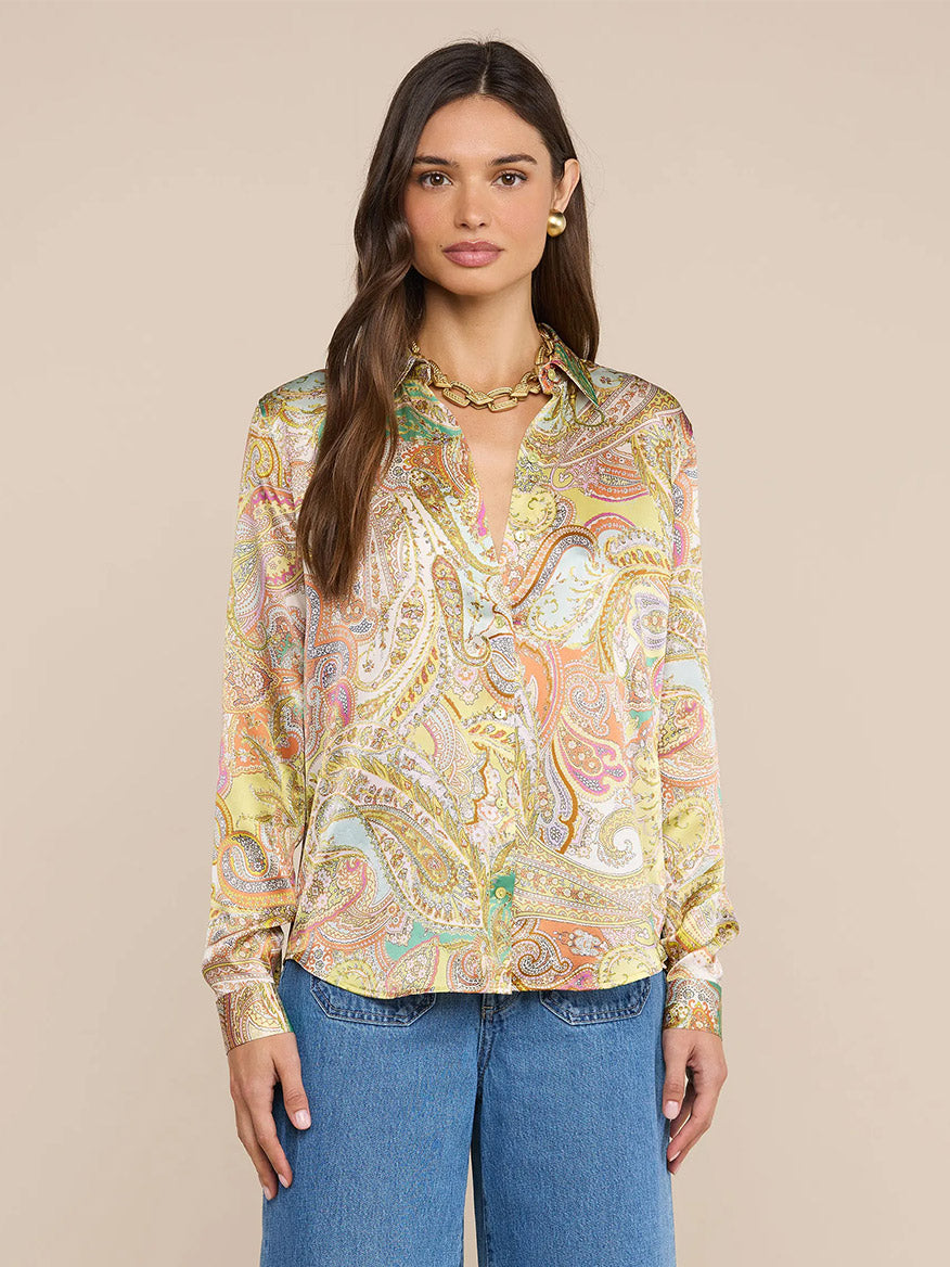 A woman stands against a beige background, wearing the L'Agence Tyler Blouse in Green Multi Cluster Paisley, styled with blue jeans, gold earrings, and a gold chain necklace. She has long brown hair.