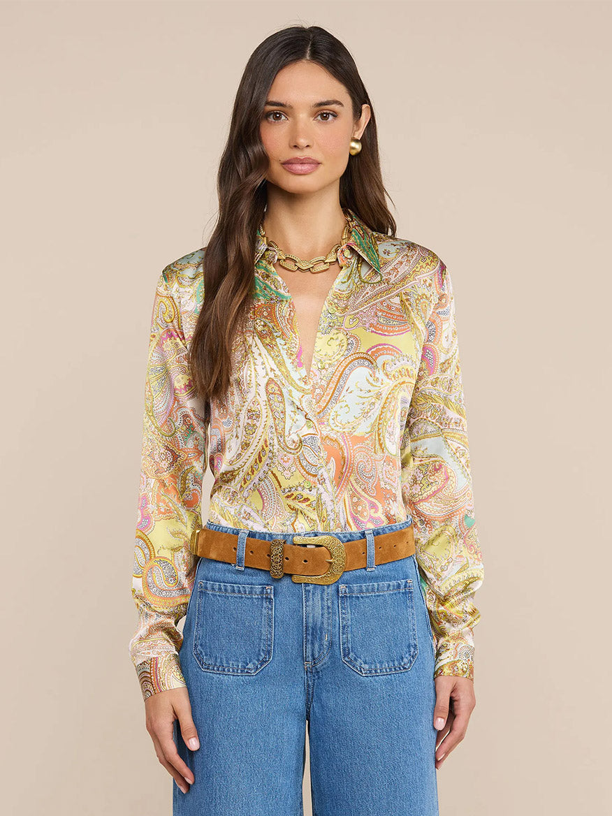 A woman wearing a L'Agence Tyler Blouse in Green Multi Cluster Paisley, along with blue high-waisted jeans, a tan belt, and a gold chain necklace, stands in front of a plain beige background.