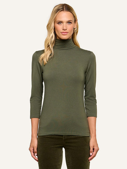 A woman stands against a plain light background wearing the L’Agence Aja Turtleneck in Army—an olive green, fitted top with three-quarter sleeves—paired with matching ultra-soft knit pants.
