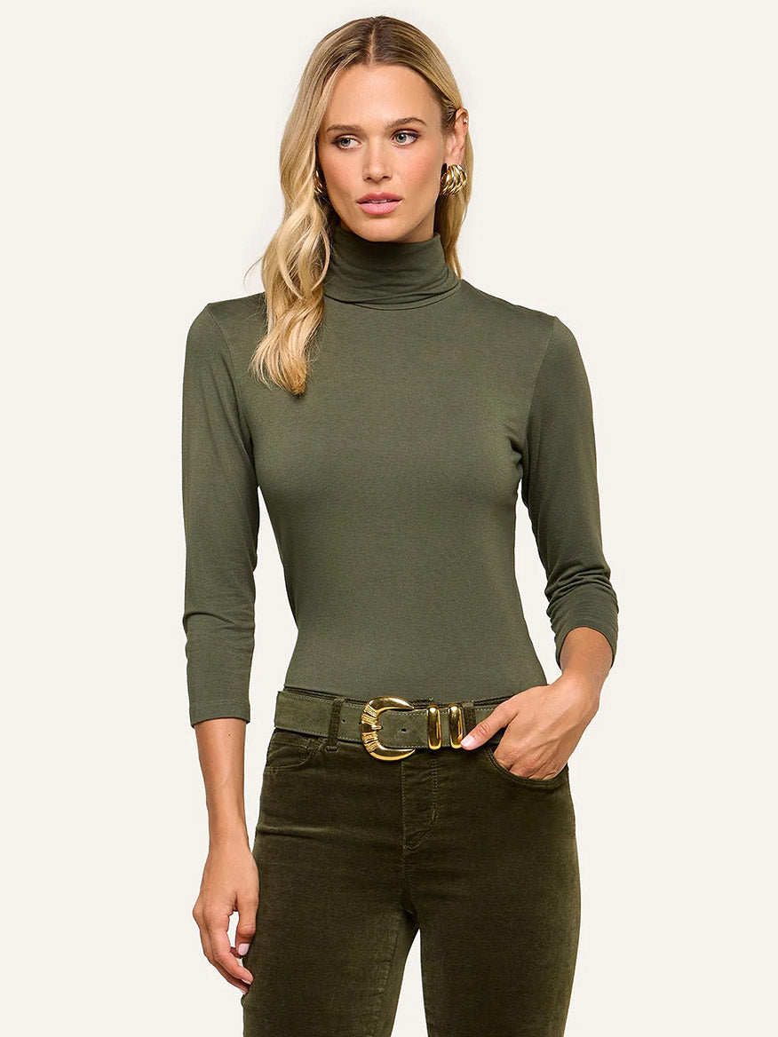 Woman wearing the L’Agence Aja Turtleneck in Army—an ultra-soft olive green knit top with three-quarter sleeves—paired with matching olive green pants, styled with a gold belt and gold hoop earrings for a chic look.