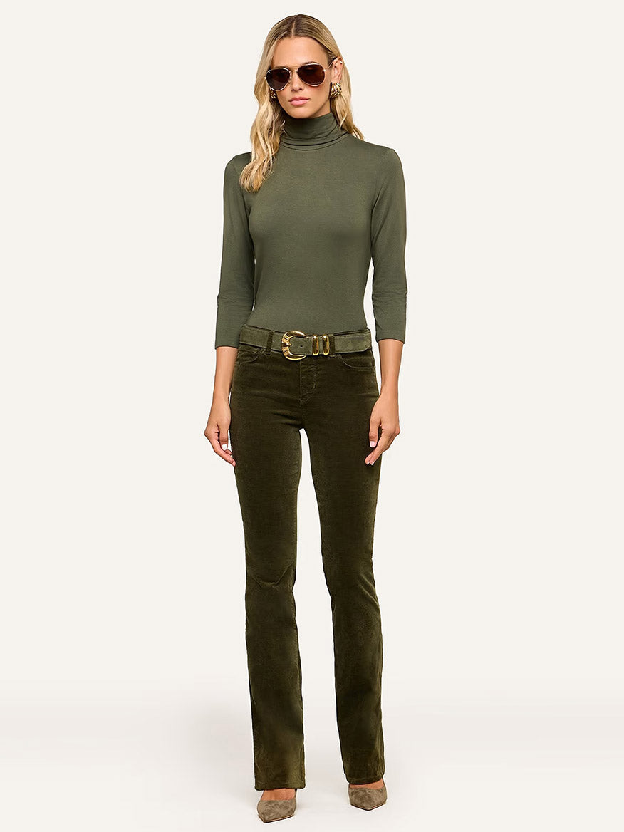 A woman in sunglasses stands against a neutral background, wearing the L’Agence Aja Turtleneck in Army paired with olive corduroy pants, a wide belt, and taupe shoes—her ultra-soft knit ensemble exudes effortless sophistication.
