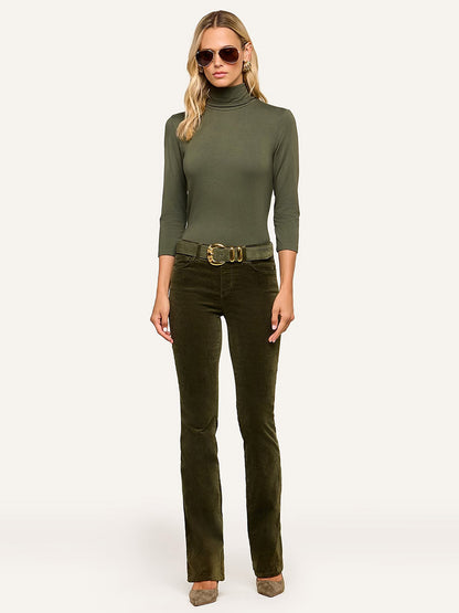 A woman in sunglasses stands against a neutral background, wearing the L’Agence Aja Turtleneck in Army paired with olive corduroy pants, a wide belt, and taupe shoes—her ultra-soft knit ensemble exudes effortless sophistication.