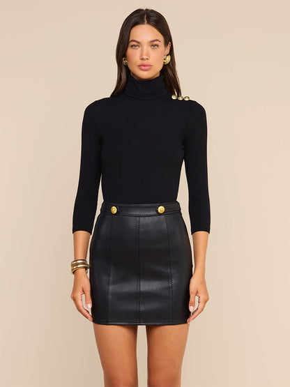 A woman wearing the L’Agence Aja Turtleneck in Black/Gold paired with a black leather mini skirt, standing against a beige background.