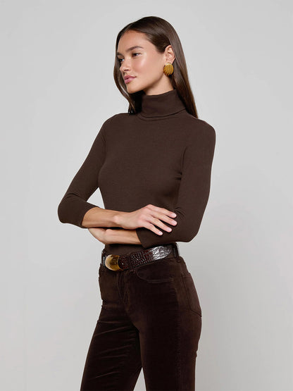 A woman wears the L'Agence Aja Turtleneck in Coffee Bean, paired with brown pants and a gold-buckle belt, standing against a plain light gray background—perfect for easy layering.