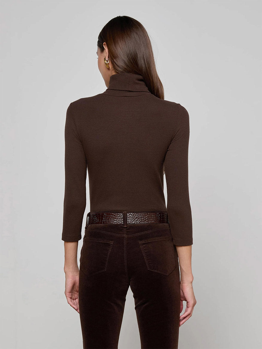 A woman with long brown hair stands with her back to the camera, showcasing the L'Agence Aja Turtleneck in Coffee Bean and dark brown belted pants for an effortlessly chic layered look against a plain background.
